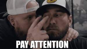 GIF: a man points two fingers at another man's eyes before pointing away from them. The caption reads "pay attention"